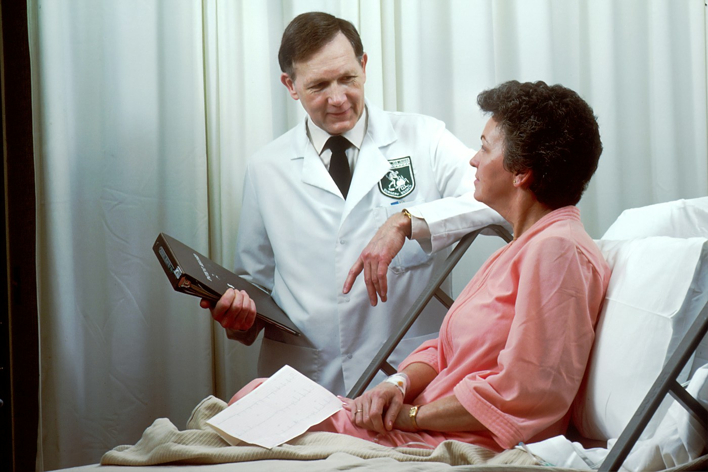 Médecin auprès d’une patiente dans un service hospitalier. Illustration du dialogue de suivi, sans diagnostic attribué.